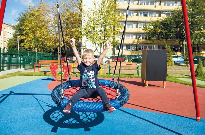 Chlapec s blond vlasmi a modrým tričkom sedí na hojdačke v tvare hniezda na ihrisku. Má zdvihnuté ruky a usmieva sa. Ihrisko je farebné s modrým a červeným gumovým povrchom, obklopené lavičkami, zeleňou a oplotením. V pozadí sú bytové domy a stromy.